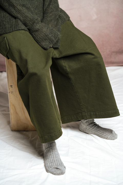 Angled view of model sitting on wooden box. Model wearing Mónica Cordera chunky cotton pocket pant in green. Paired with Mónica Cordera soft alpaca turtleneck in green